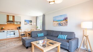 A modern living room with a grey sofa, a wooden coffee table, and a kitchen area with white cabinets and a stove. - Meerliebe - Stern FeWo Strandstraße (Karlshagen)