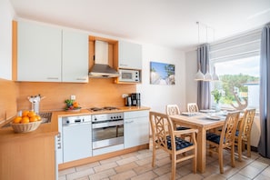A compact kitchen with a microwave, stove, and sink. There is a dining table with chairs and a window with curtains.