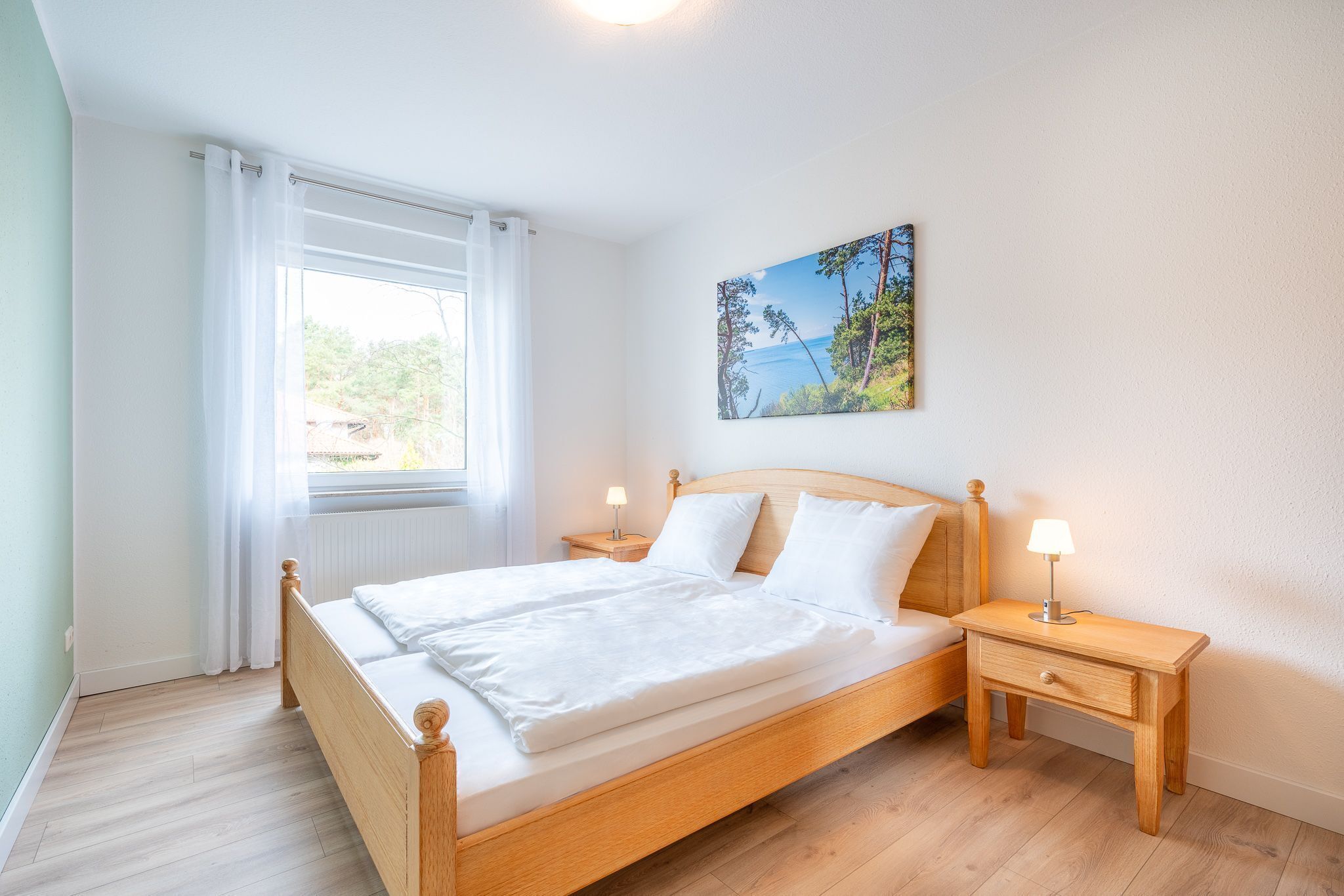 Ein Schlafzimmer mit einem hölzernen Bett, weißer Bettwäsche, einem Nachttisch mit Lampe, einem Fenster mit Vorhängen und einem Bild an der Wand.