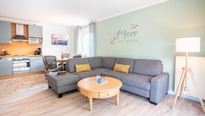 Ein modernes Wohnzimmer mit einem grauen Ecksofa, einem Holztisch und einem Küchenbereich im Hintergrund.