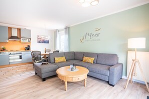 Uma sala de estar moderna com um sofá modular cinzento, uma mesa de centro de madeira e uma área de cozinha ao fundo.