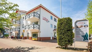 A multi-story building with a white facade and red accents, featuring balconies and a signboard with text. - Seewind - Stern FeWo Strandstrasse (Karlshagen)