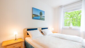 A double bed with white linens, a nightstand with a lamp, and a painting of a lake scene on the wall. - Seewind - Stern FeWo Strandstrasse (Karlshagen)