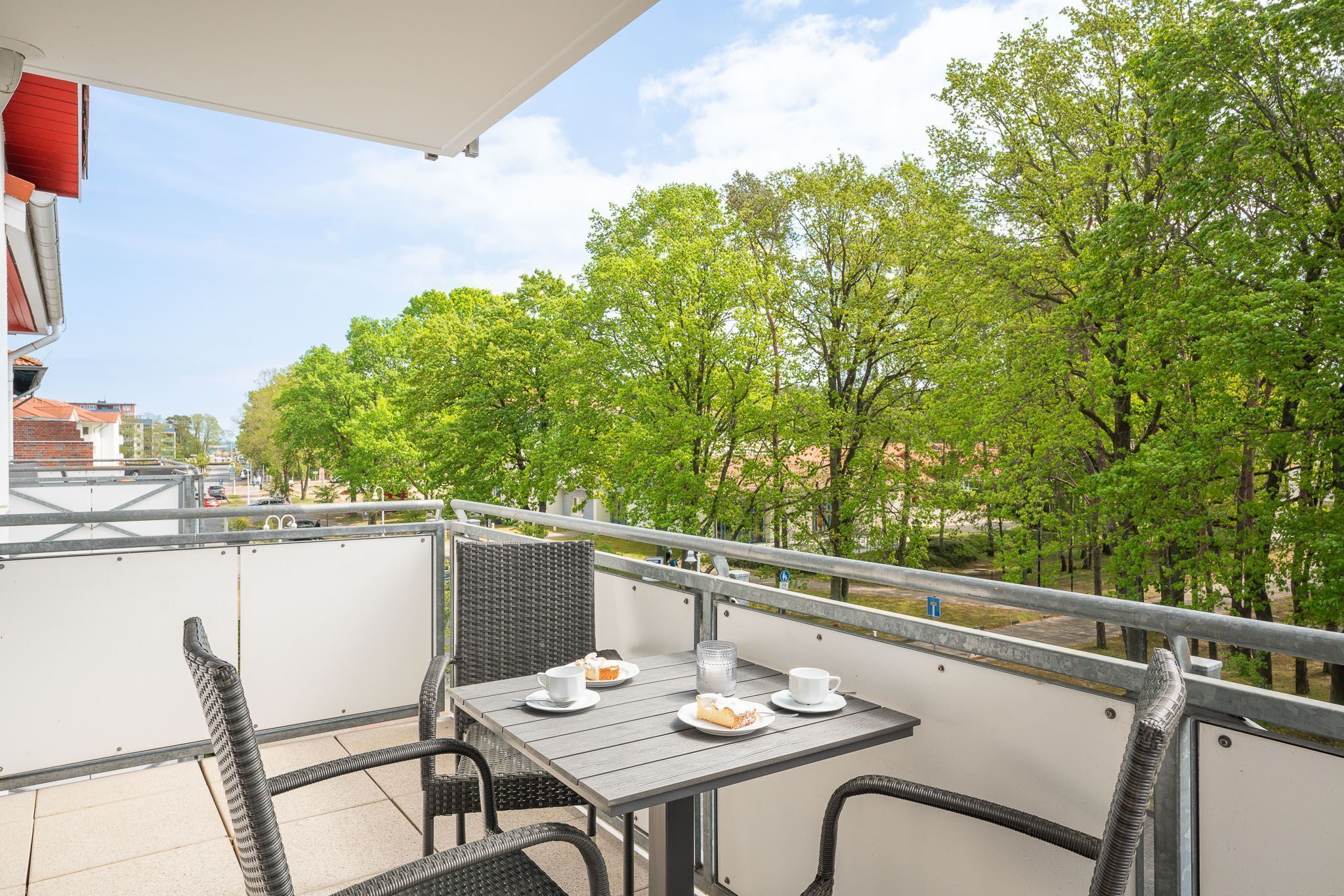 A balcony with a table set for two, surrounded by wicker chairs, overlooking a tree-lined street.