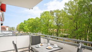 A balcony with a table set for two, surrounded by wicker chairs, overlooking a tree-lined street. - Seewind - Stern FeWo Strandstrasse (Karlshagen)