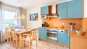 Een kleine keuken met blauwgroene kastjes, een houten eettafel en uitzicht op zee.