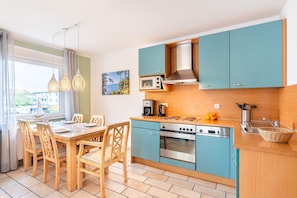 A compact kitchen with teal cabinets, a wooden dining table, and a view of the sea through the window.