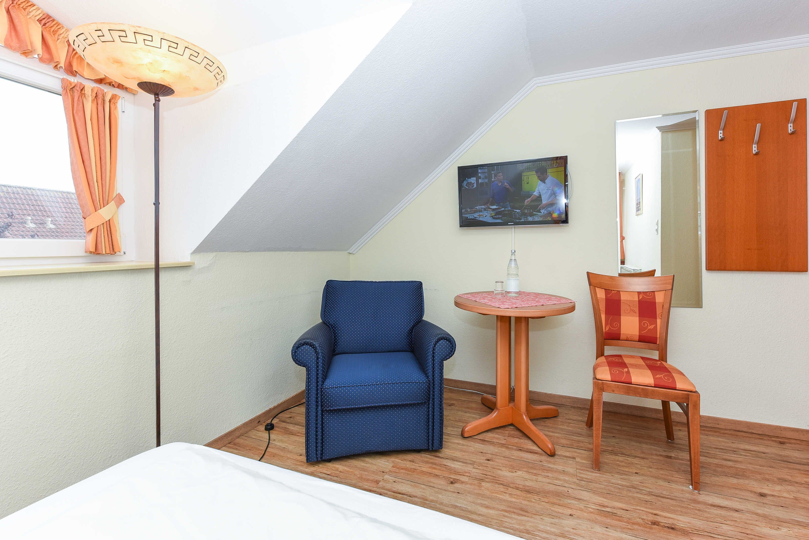 A room with a sloped ceiling, a bed, a blue armchair, a wooden chair, a small round table, and a TV mounted on the wall.