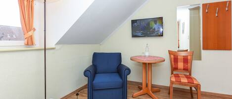 A room with a sloped ceiling, a bed, a blue armchair, a wooden chair, a small round table, and a TV mounted on the wall.