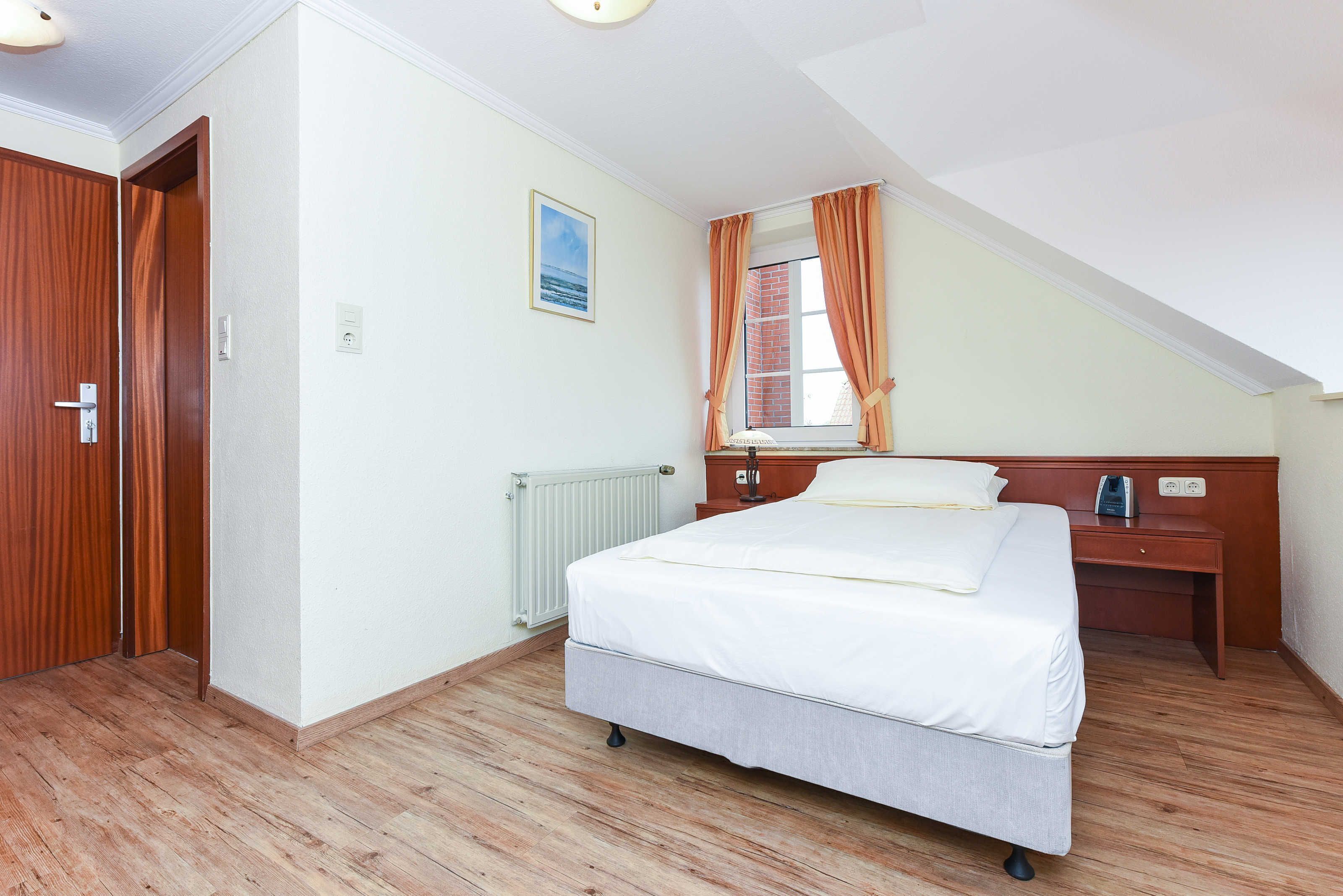 A bedroom with a bed, a nightstand, a window with curtains, a radiator, and a door.