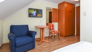 Une pièce comprenant un lit, un fauteuil bleu, une armoire en bois, une petite table avec une chaise et un téléviseur fixé au mur.