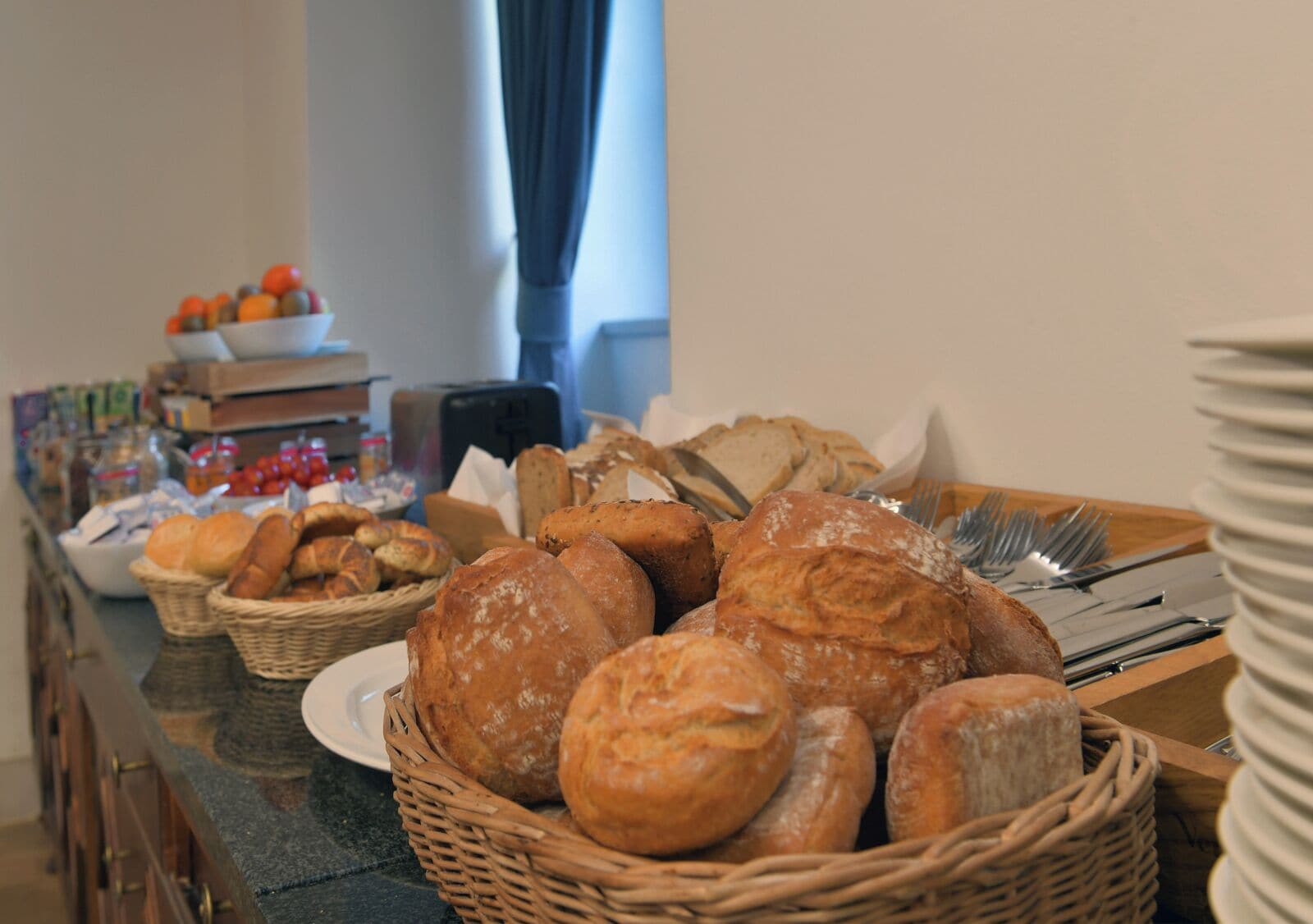 A buffet table with various breads and pastries, a bowl of tomatoes, and condiments.