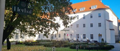 A white building with a red roof, labeled "Herbal Hotel," surrounded by a garden with a cobblestone path.