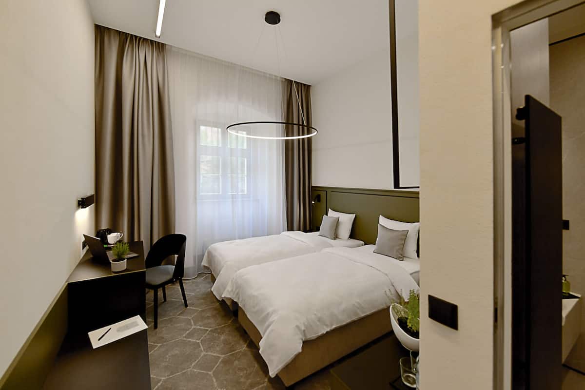 A hotel room with two beds, a desk with a laptop, a chair, a desk lamp, a potted plant, and a window with curtains.