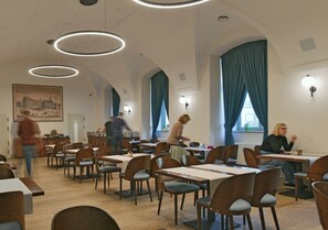 Un comedor amplio con varias mesas y sillas, techos altos con lámparas circulares y ventanas grandes con cortinas.