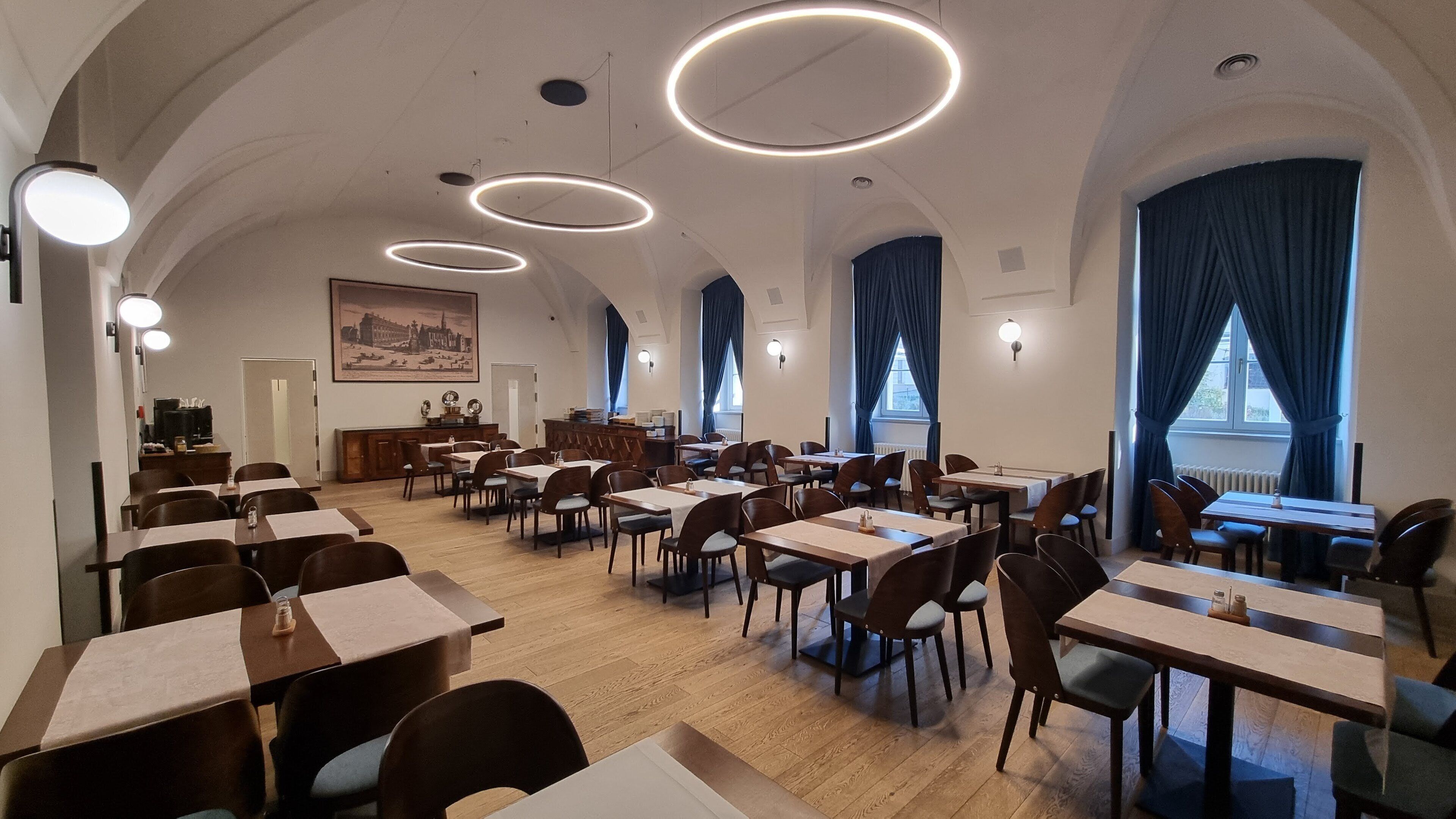 A spacious dining area with multiple tables and chairs, a central counter, and large windows with blue curtains.