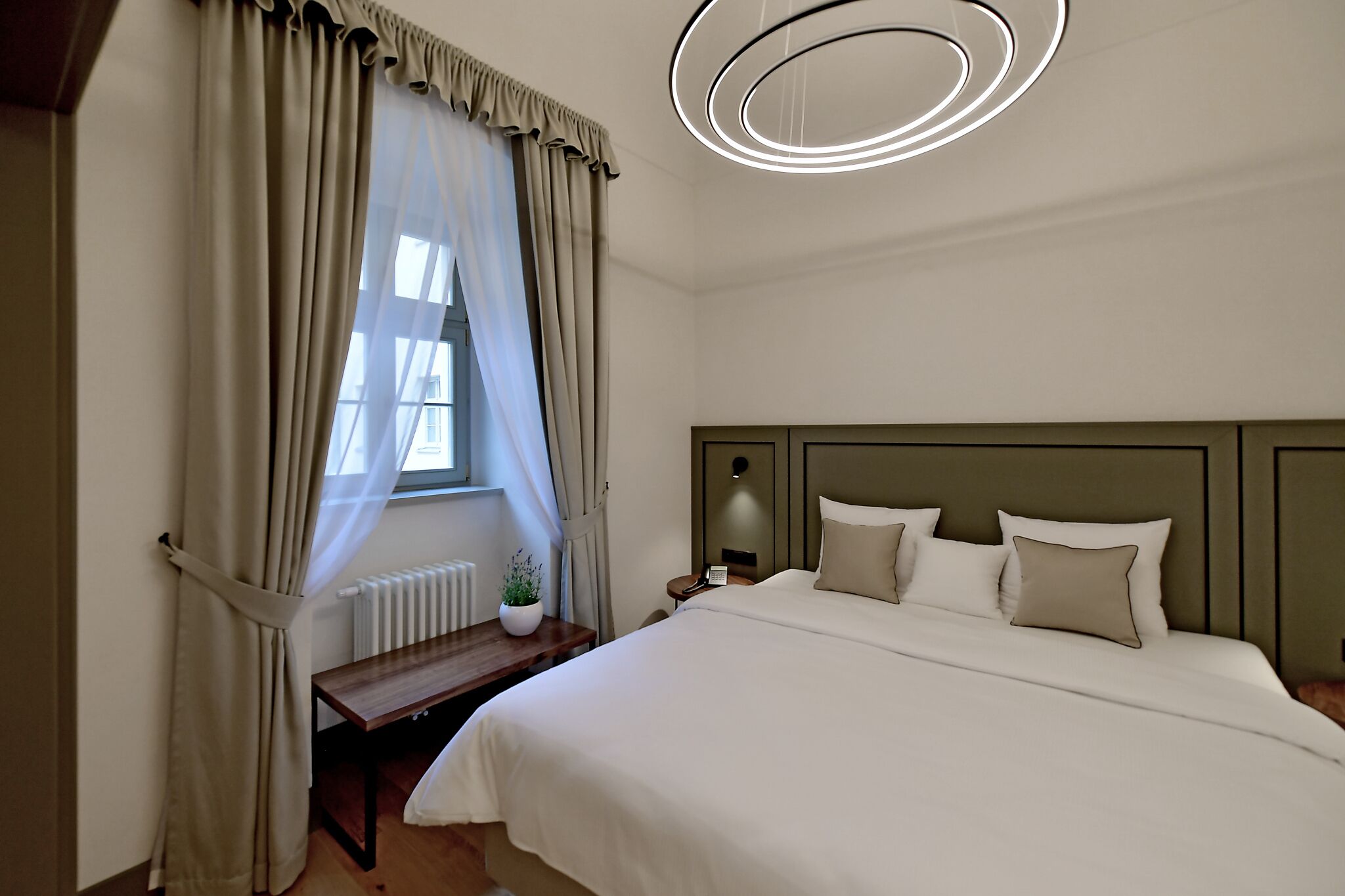 A bedroom with a large bed, a bedside table with a lamp, a window with curtains, and a modern ceiling light fixture.