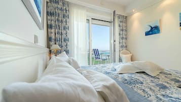 A hotel room with a large bed, a view of the sea, a chair by the window, and a painting on the wall.