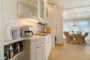 A modern kitchen with white cabinetry, a coffee maker, a kettle, and a dining area with wicker chairs and a chandelier.