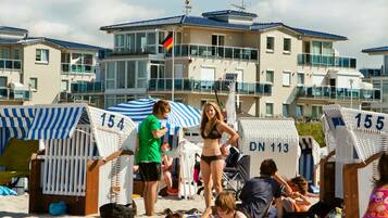 Hébergement en bord de mer avec des cabanes numérotées sur la plage, un drapeau et un bâtiment en arrière-plan.