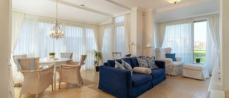 Amplia sala de estar con un sofá azul, sillas de mimbre, una mesa de comedor y ventanas grandes con cortinas translúcidas.