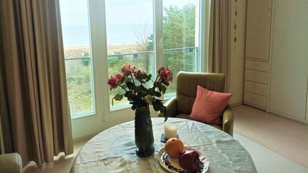Ein Zimmer mit großem Fenster, einem Tisch mit Blumen, einer Kerze und einem Teller mit Obst.