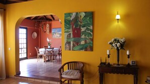 Quarto com parede amarela, uma cadeira de madeira, uma mesa com cadeiras, um quadro e um abajur.
