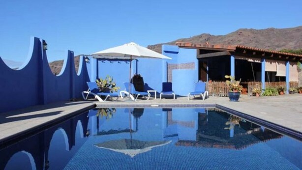 Piscina azul cristalina com espreguiçadeiras e guarda-sóis, cercada por um edifício azul e montanhas ao fundo.