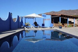 A clear blue swimming pool with lounge chairs and umbrellas, surrounded by a blue building with a mountain backdrop. - Villa Greca, Spanien (Barros)