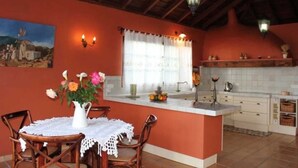 Uma cozinha com ilha central, cadeiras de madeira, mesa de jantar com toalha rendada e um quadro na parede.