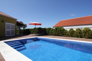 Een zwembad met treden die erin leiden, omringd door een terras met een rode parasol en lounge stoelen.