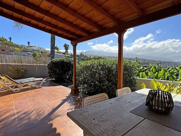 Une terrasse couverte, dotée d’une table et de chaises, entourée de verdure et offrant une vue sur le paysage urbain.