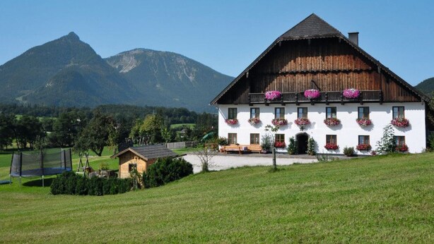 Un edificio bianco con persiane di legno, finestre con fioriere e una montagna sullo sfondo.