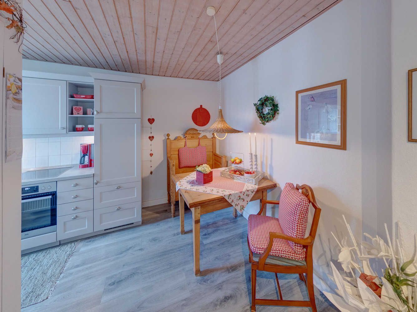 Una habitación acogedora con techo de madera, una mesa de comedor con mantel a cuadros, una silla de madera y una zona de cocina con armarios blancos y un electrodoméstico rojo.