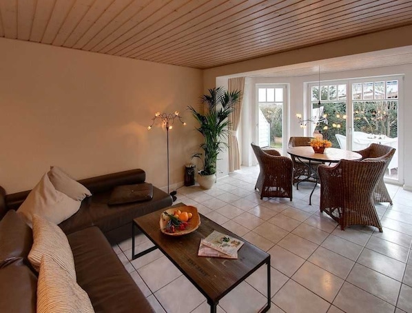 A living room with a sofa, coffee table, and dining area with wicker chairs. There is a potted plant and a floor lamp.