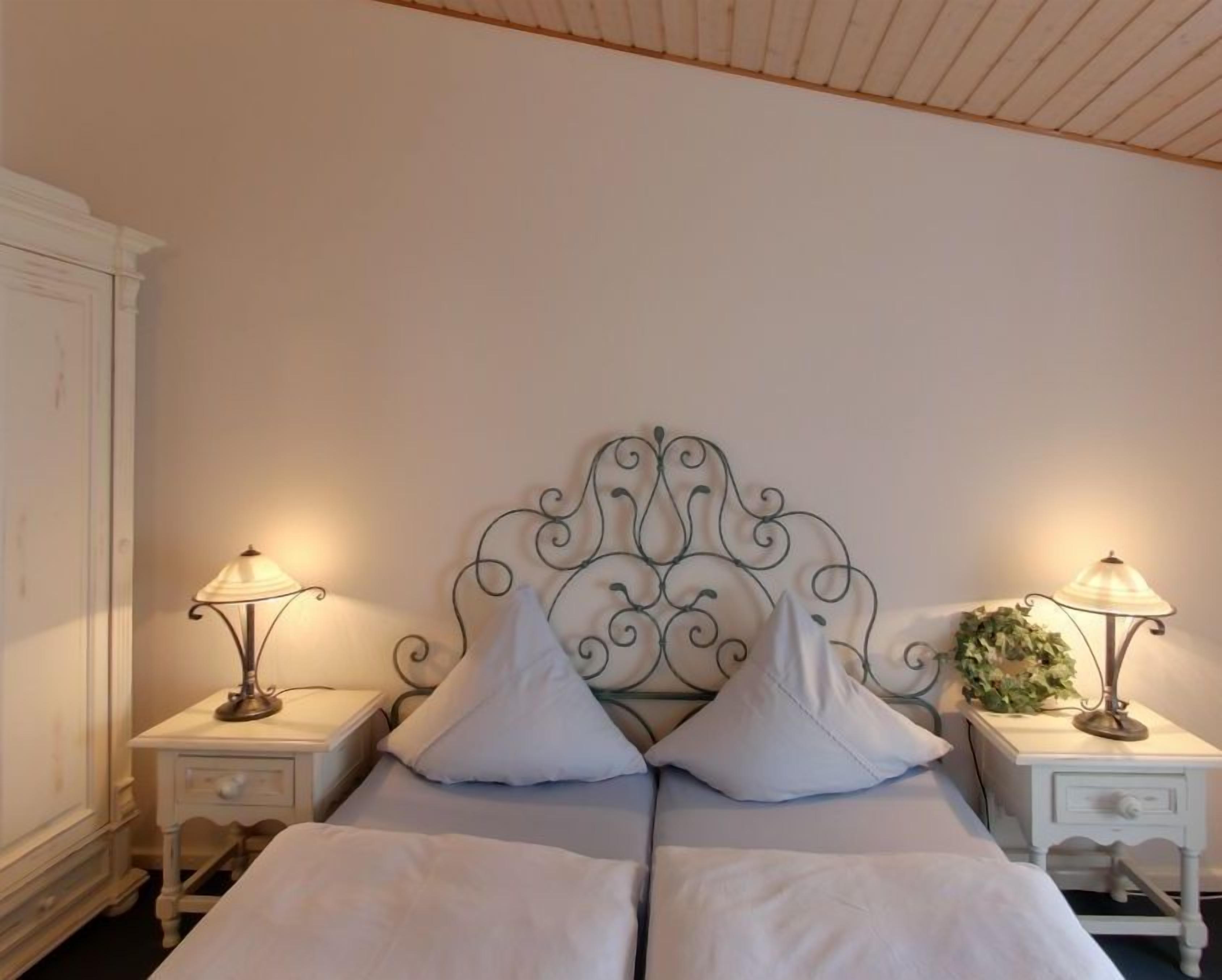 Una cama doble con un cabecero metálico decorativo, dos mesitas de noche con lámparas y un armario blanco.