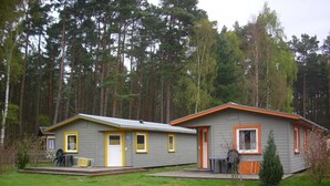 Twee vakantiehuisjes met grijze buitenmuren en gele accenten, omgeven door een bos. Elk huisje heeft een deur en ramen, en één heeft een veranda met een tafel en stoelen.