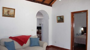 Una sala de estar con un sofá color beige, cojines rojos y azules, una mesa de centro de madera y una pequeña cesta. Hay dos cuadros enmarcados en las paredes y un umbral que da a otra habitación.