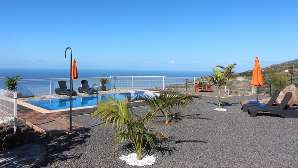 Zona de piscina con sillones de descanso y sombrillas, vista al mar y piscina cercada.
