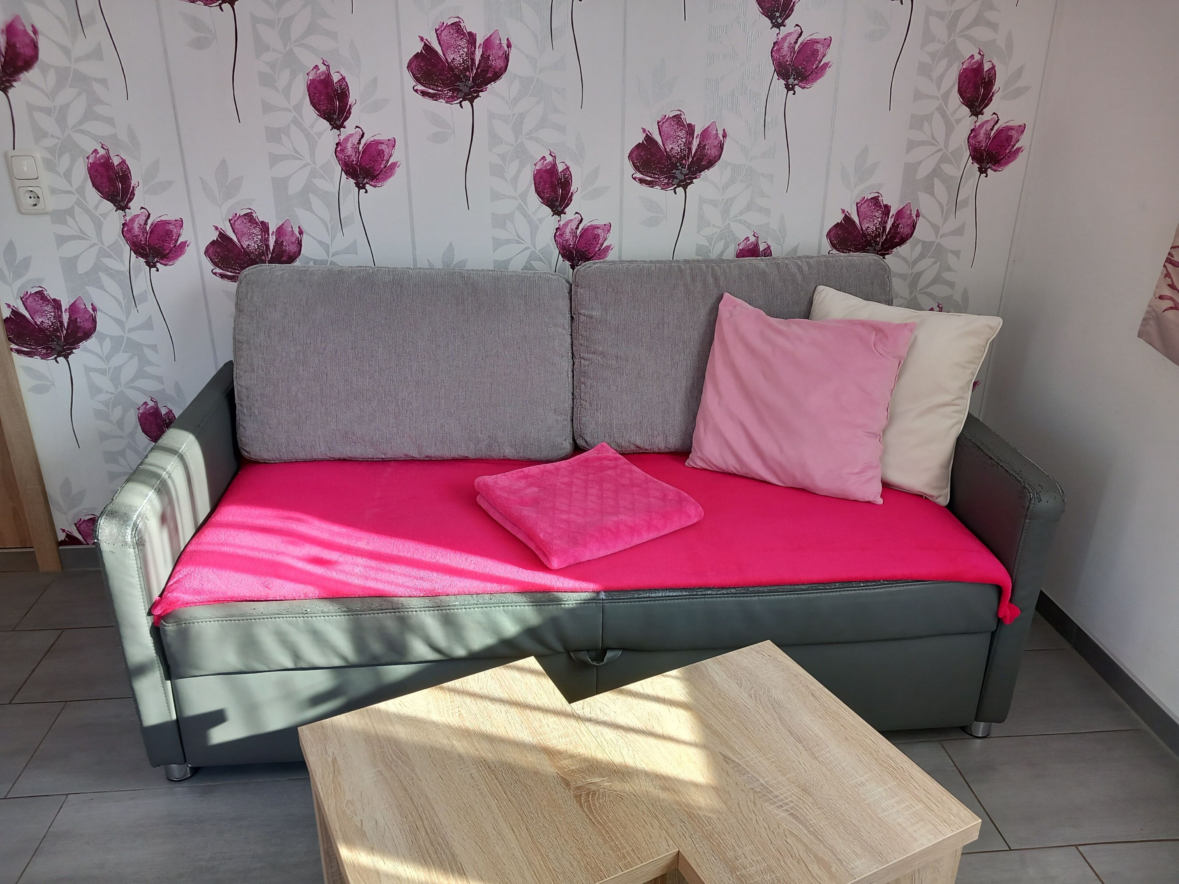 Un sofá gris con una funda rosa brillante y dos almohadas, una mesa de centro de madera al frente y una pared con estampado floral.