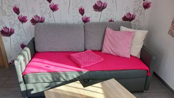 Un sofá gris con una funda rosa brillante y dos almohadas, una mesa de centro de madera al frente y una pared con estampado floral.