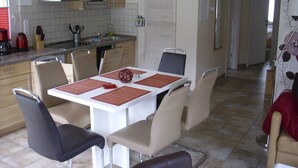 A modern kitchen with a dining area, featuring a white table with red placemats, beige and black chairs, and a microwave above the sink. - Ferienhaus L 04 - Ferienhaus 4 (Trassenheide)