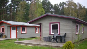 A two-story cabin with a red-framed window and a small porch area with plastic chairs. - Ferienhaus L 04 - Ferienhaus 4 (Trassenheide)