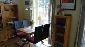 Un coin repas avec une table, des chaises et une lampe. Il y a une bibliothèque sur la gauche et une fenêtre avec des rideaux.