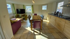 A compact living space with a dining table, chairs, a sofa, a TV, and a kitchen area with cabinets and a window.