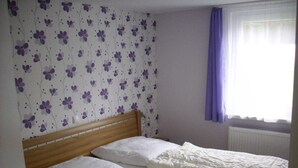 A bedroom with a wooden headboard, two single beds, white bedding, a floral wallpaper pattern, and a window with purple curtains.