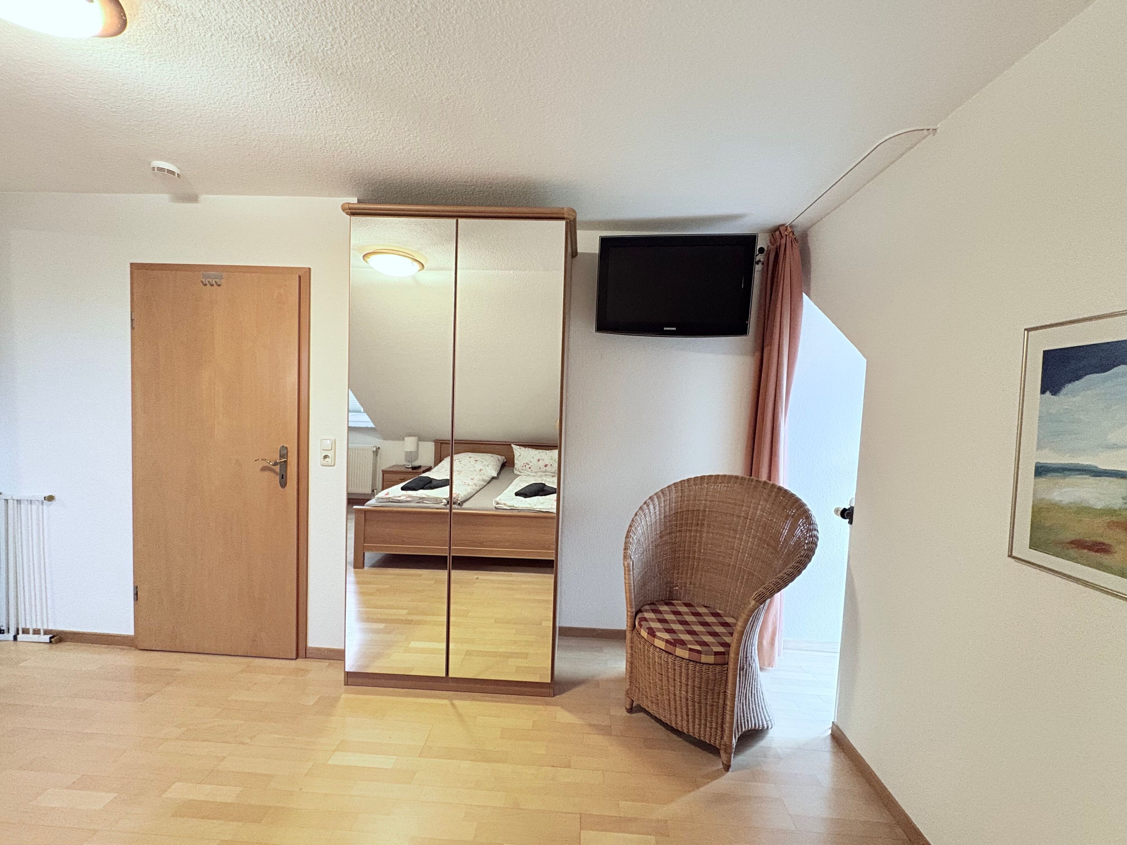 A bedroom with a wooden bed, a mirror wardrobe, a wicker chair, a flat-screen TV, and a painting on the wall.