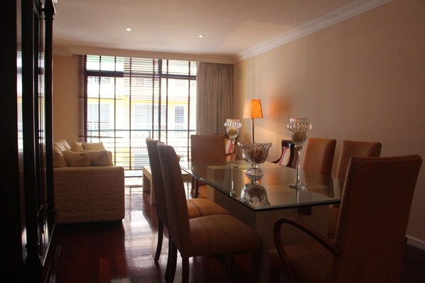 A hotel room with a glass dining table, four chairs, a sofa, and a large window with curtains.