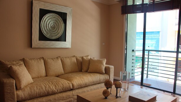 Ein beiges Sofa mit einem großen abstrakten Gemälde darüber, ein Couchtisch aus Holz mit Dekorationselementen und Blick auf ein Gebäude durch eine Glastür.