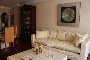 A living room with a beige sofa, a coffee table with decorative items, a wicker chair, and a cabinet with glassware.